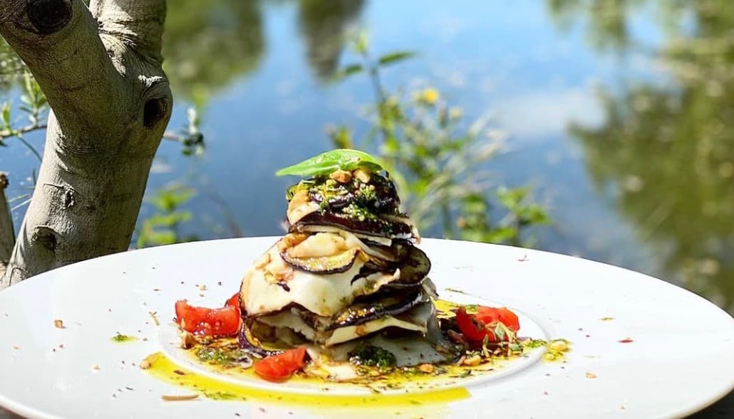 Türmchen aus gegrillter Aubergine und Mozzarella auf weißem Teller, dahinter der Olivenbaum und das Wasser
