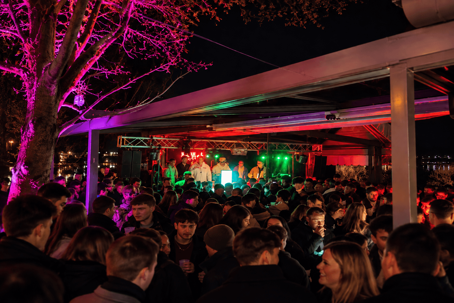 Open-Air-Bühne unter dem Dach am Wasser bei Nacht, pink beleuchteter Baum und dicht gedrängte Gäste vor den DJs