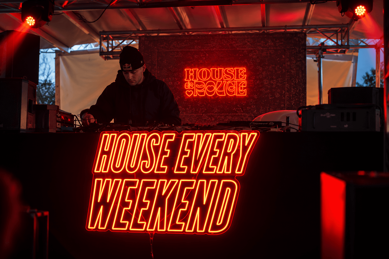 DJ am Pult hinter der Neonschrift „House Every Weekend“, darüber „House de Rouge“